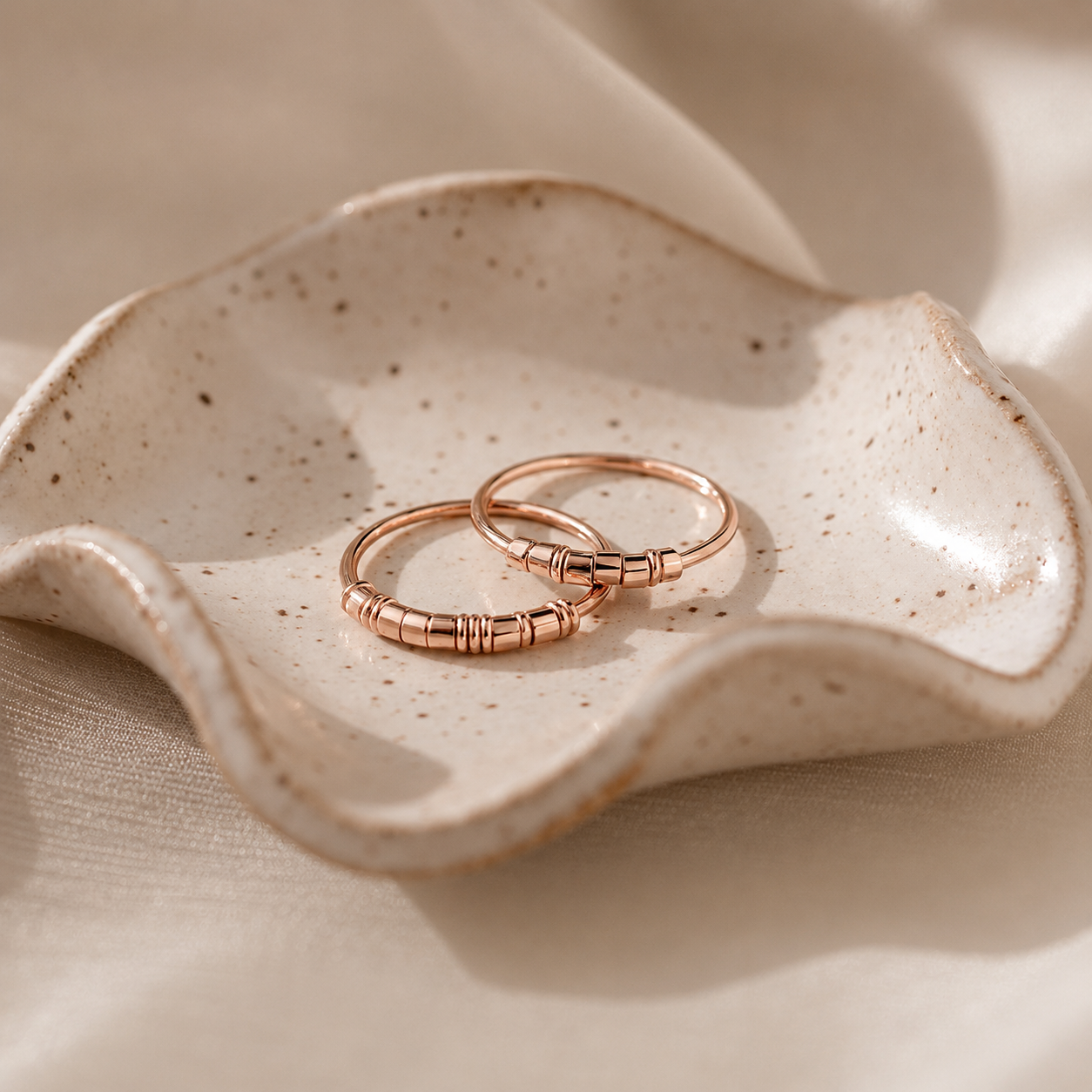 Two rose gold rings on a textured ceramic dish with a beige fabric background